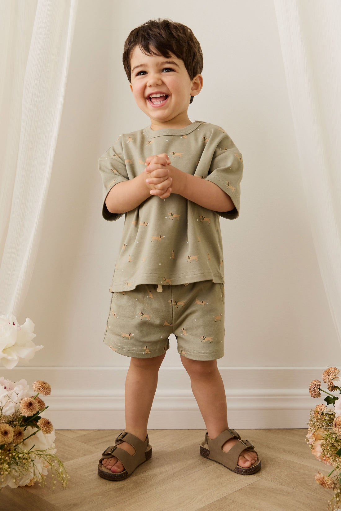Child wearing a matching outfit with a light green shirt and shorts, standing against a white curtain background.