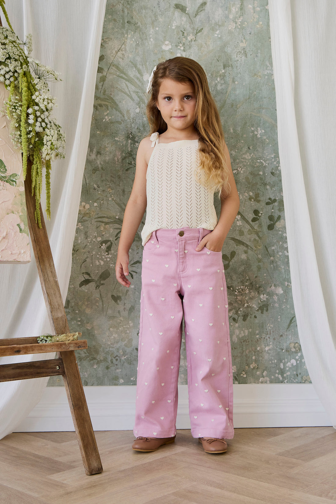 Young girl wearing a white top and pink pants with a floral pattern in a room with a decorative wall.