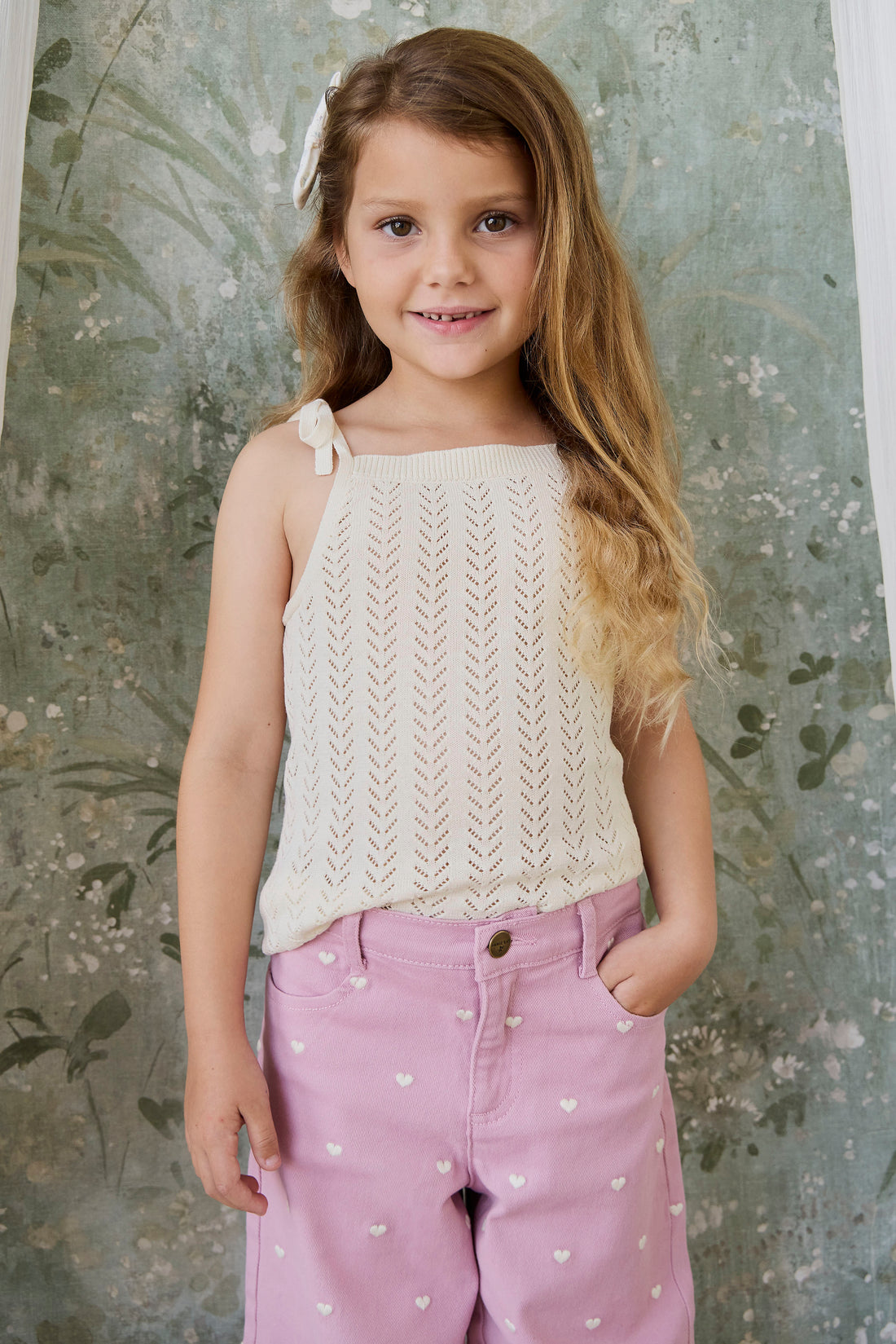 A young girl with long light brown hair stands against a textured, vintage-style green floral backdrop. She is wearing a cream-colored sleeveless knit top with a small bow on the shoulder and pink pants decorated with small white hearts.
