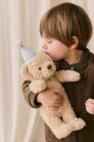 Child holding a teddy bear with a party hat against a neutral background