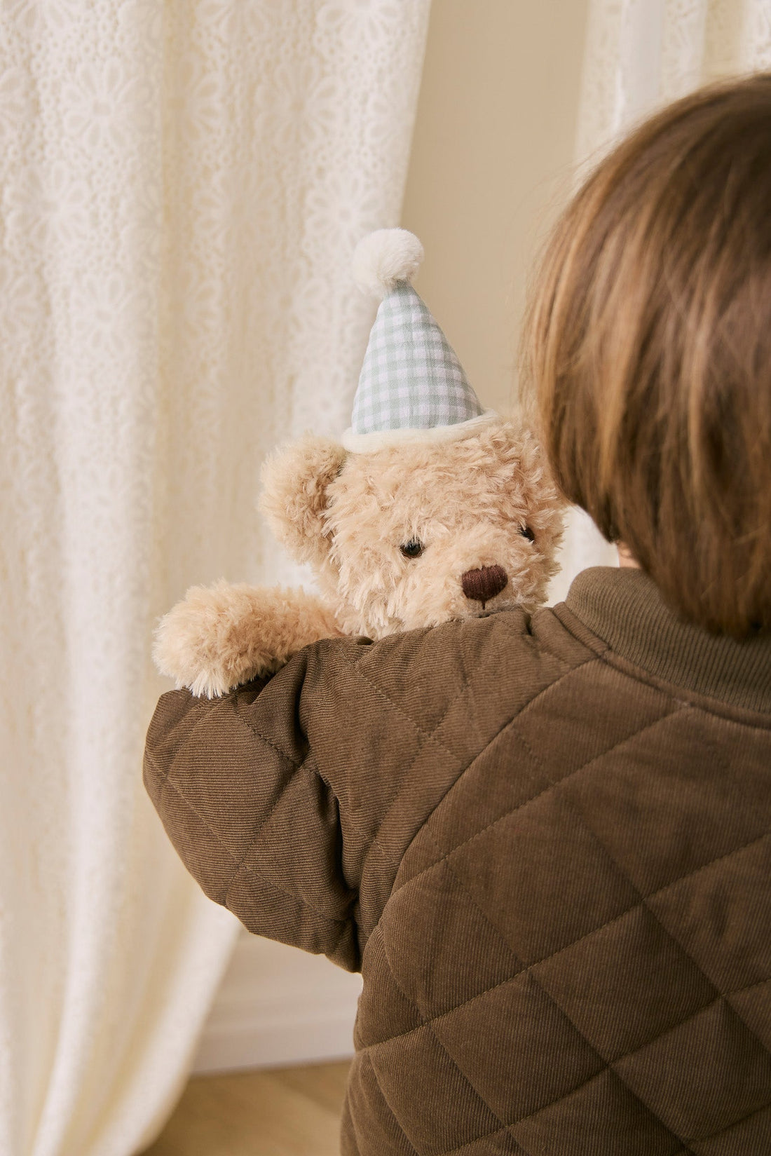 Person holding a teddy bear wearing a party hat in front of a curtain