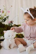 A toddler wearing a pink knit cardigan and white bunny-ear headband sits beside a fluffy white plush kitty toy.