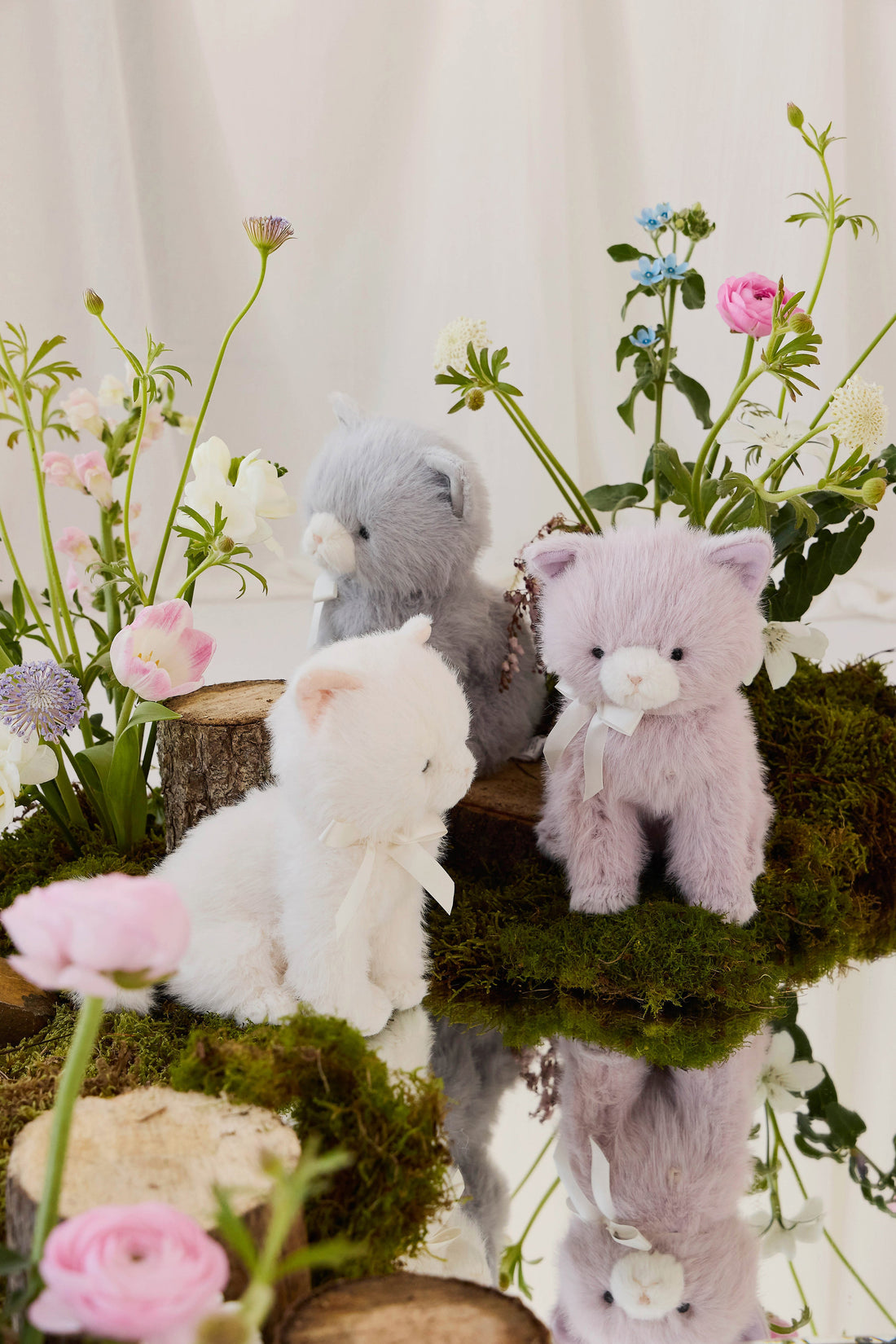Three Jamie Kay plush kitty toys—one violet with a white satin bow, one grey-blue, and one white—seated and standing together outdoors on a mossy log near a pond surrounded by pink and white flowers.