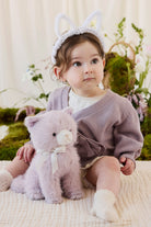 A toddler wearing a knit violet cardigan and white bunny-ear headband cuddling a soft violet-toned plush kitty toy by Jamie Kay.