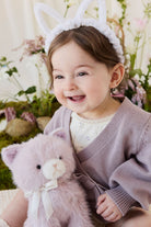 A toddler wearing a knit violet cardigan and white bunny-ear headband cuddling a soft violet-toned plush kitty toy by Jamie Kay.
