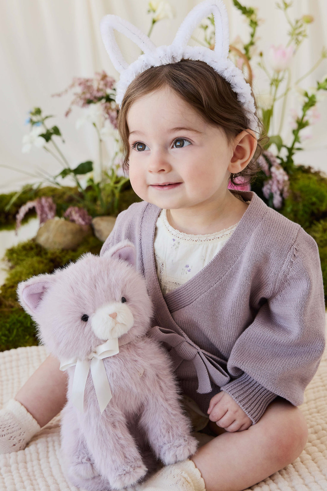 A toddler wearing a knit violet cardigan and white bunny-ear headband cuddling a soft violet-toned plush kitty toy by Jamie Kay.