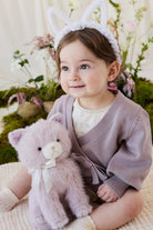 A toddler wearing a knit violet cardigan and white bunny-ear headband cuddling a soft violet-toned plush kitty toy by Jamie Kay.