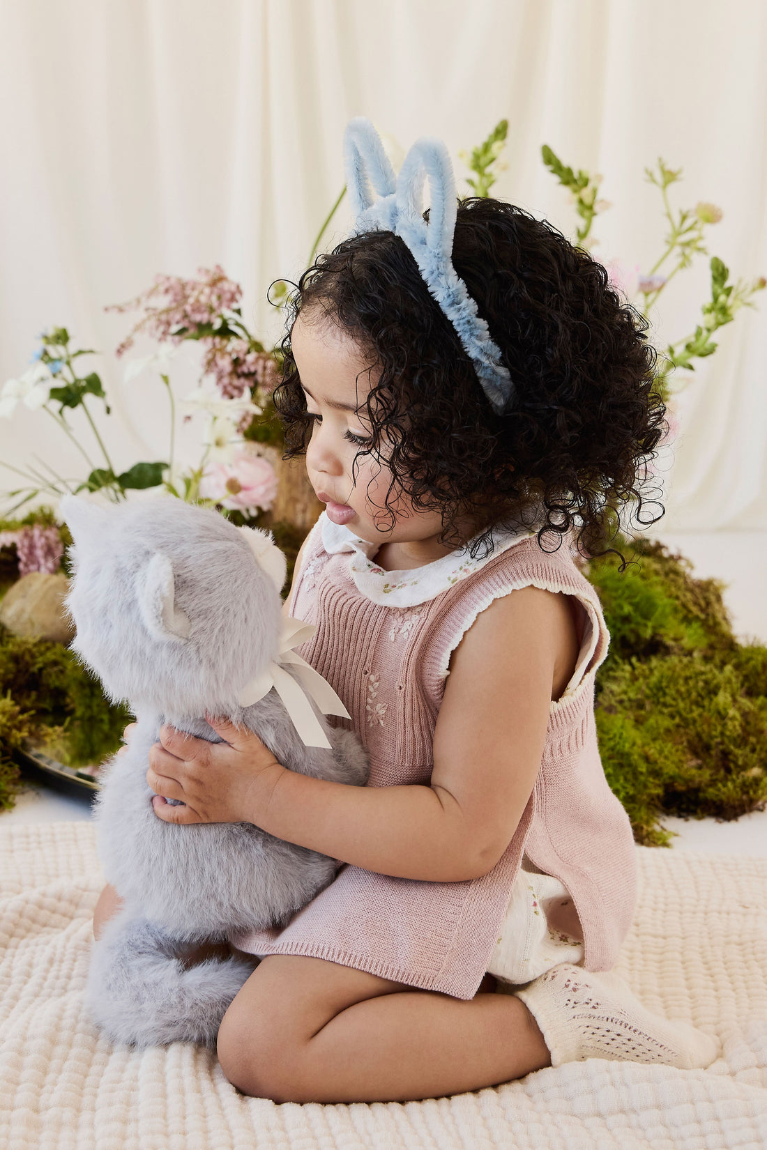 A child wearing a blue headband with bunny ears, a pink knit vest, and white shorts, kneeling on a blanket while holding the soft grey plush kitten toy.