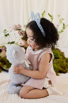 A child wearing a blue headband with bunny ears, a pink knit vest, and white shorts, kneeling on a blanket while holding the soft grey plush kitten toy.