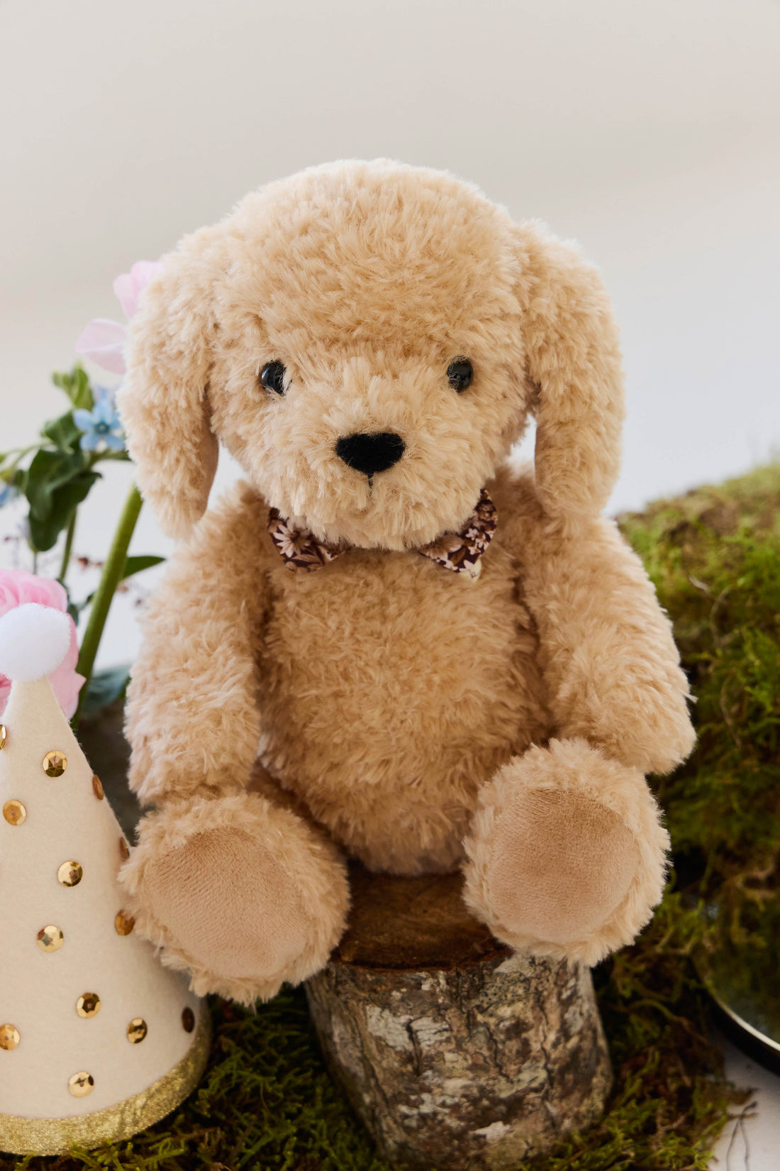 An eye-level studio product shot of the "Snuggle Bunnies - Honey the Puppy by Jamie Kay" plush toy sitting upright on a small wooden log stump. The puppy is made of soft, textured honey-toned faux fur with long floppy ears, shiny dark brown button eyes, a stitched dark brown nose, and a checkered ribbon collar. The rustic setting is styled with patches of green moss at the base and a party hat with gold foil polka dots positioned beside the toy.