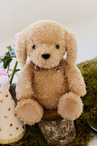 An eye-level studio product shot of the "Snuggle Bunnies - Honey the Puppy by Jamie Kay" plush toy sitting upright on a small wooden log stump. The puppy is made of soft, textured honey-toned faux fur with long floppy ears, shiny dark brown button eyes, a stitched dark brown nose, and a checkered ribbon collar. The rustic setting is styled with patches of green moss at the base and a party hat with gold foil polka dots positioned beside the toy.