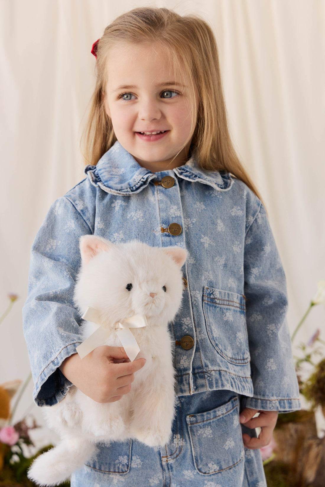 A young child with long blonde hair, wearing a denim jacket, smiles while holding a fluffy white plush kitty toy against a neutral background.