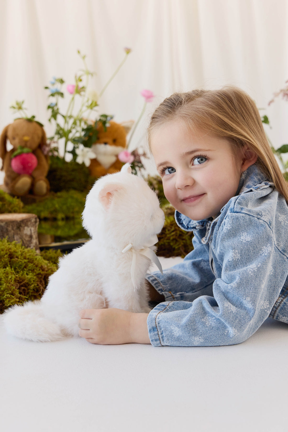 A young child with long blonde hair, wearing a denim jacket, smiles while holding a fluffy white plush kitty toy against a neutral background.