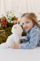 A young child with long blonde hair, wearing a denim jacket, smiles while holding a fluffy white plush kitty toy against a neutral background.