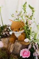 A plush toy fox sitting on a rustic wooden stump surrounded by blooming spring flowers and lush greenery.