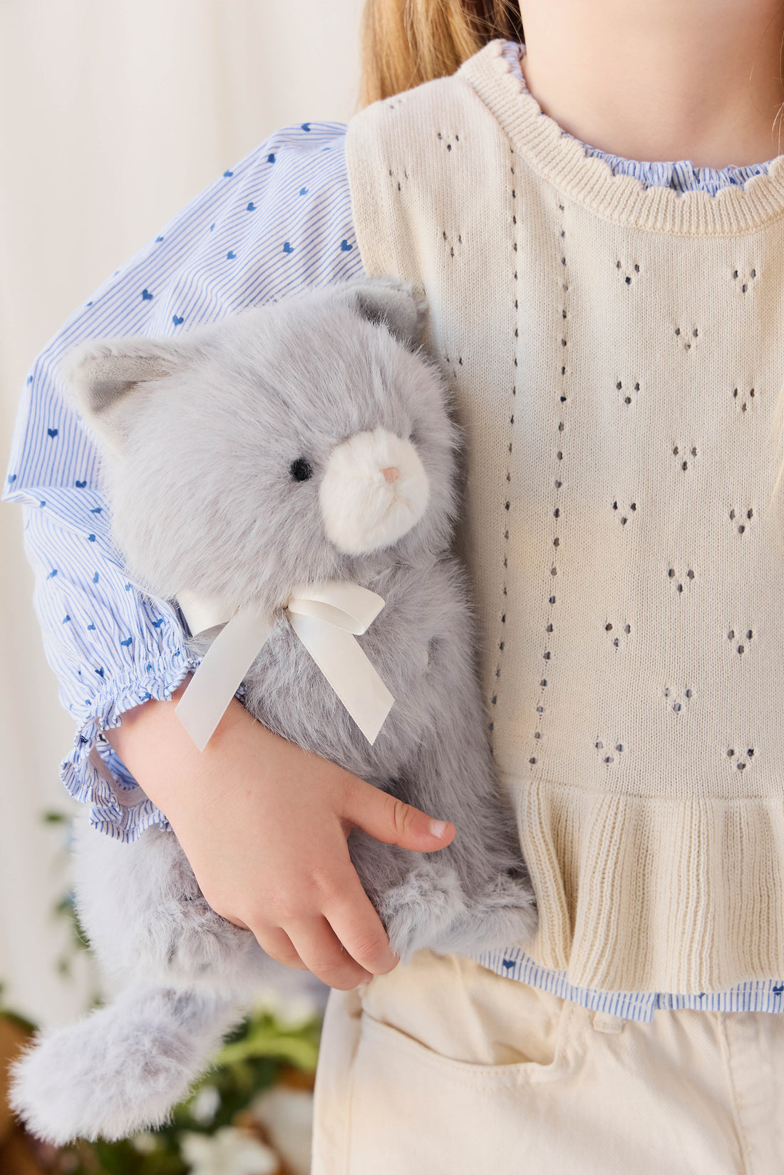 A child wearing a cream-colored knit vest and a blue patterned blouse while holding the soft grey plush kitten toy.