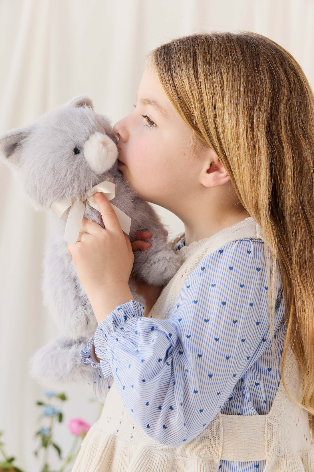 A child with long brown hair pressing her cheek gently against the soft grey plush kitten toy, wearing a cream-colored knit vest over a blue patterned blouse.