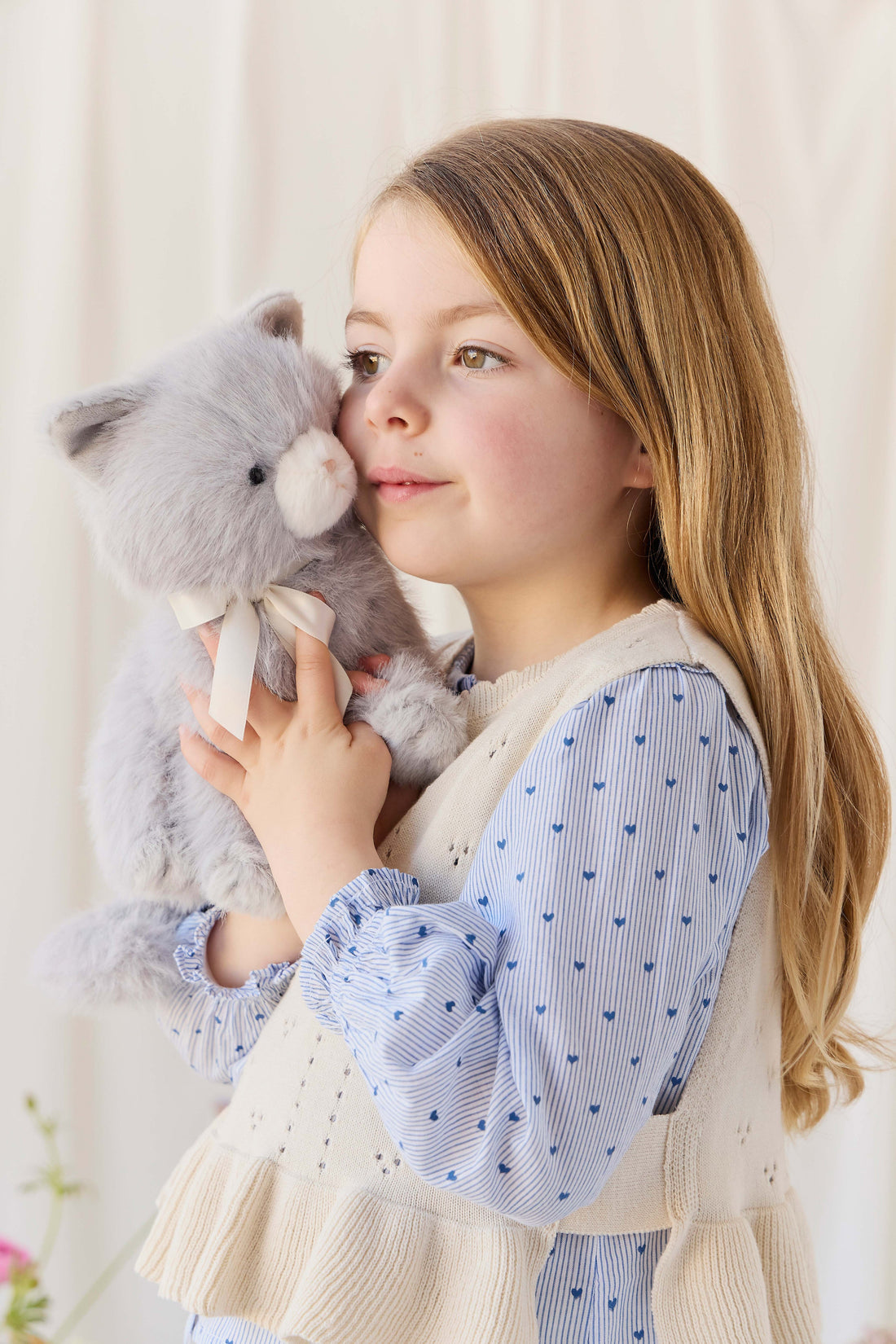 A child holding the soft grey plush kitten toy close while looking off to the side, wearing a patterned blue blouse and a cream-colored knit vest.