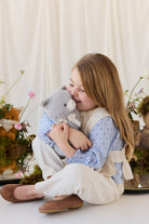 A child with long brown hair smiling as she hugs the soft grey plush kitten toy, wearing a cream-colored knit vest over a blue patterned blouse.