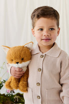 A young child smiling gently while wearing a beige knit button-down cardigan, holding a plush fox toy against a soft, neutral-toned background.