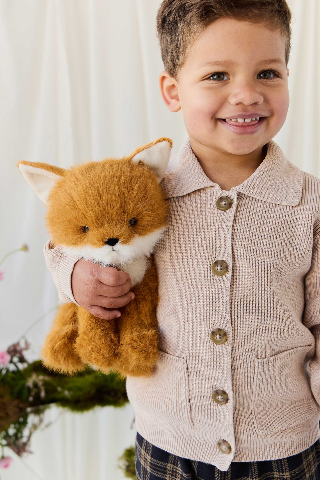 A young child smiling warmly while holding a plush fox toy, wearing a knit beige button-down collared cardigan over a plaid shirt.