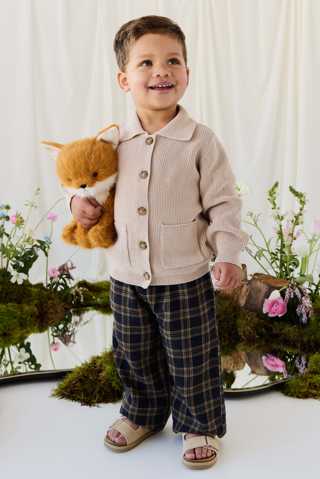 A young child smiling warmly while wearing a beige knit button-down cardigan and patterned trousers, holding a plush fox toy against a rustic woodland-themed background.