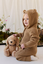 An eye-level studio portrait of a toddler sitting indoors against a light-colored backdrop, wearing a matching textured brown knit romper with an attached hood featuring small, upright bear ears and a front kangaroo pocket. Beside the toddler, the plush honey-toned puppy toy with floppy ears and a checkered ribbon collar sits upright against the backdrop. The soft, natural lighting illuminates the scene, which is styled with delicate floral arrangements and moss at the base.