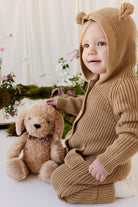An eye-level studio portrait of a toddler sitting indoors against a light-colored backdrop, wearing a matching textured brown knit romper with an attached hood featuring small, upright bear ears and a front kangaroo pocket. Beside the toddler, the plush honey-toned puppy toy with floppy ears and a checkered ribbon collar sits upright against the backdrop. The soft, natural lighting illuminates the scene, which is styled with delicate floral arrangements and moss at the base.