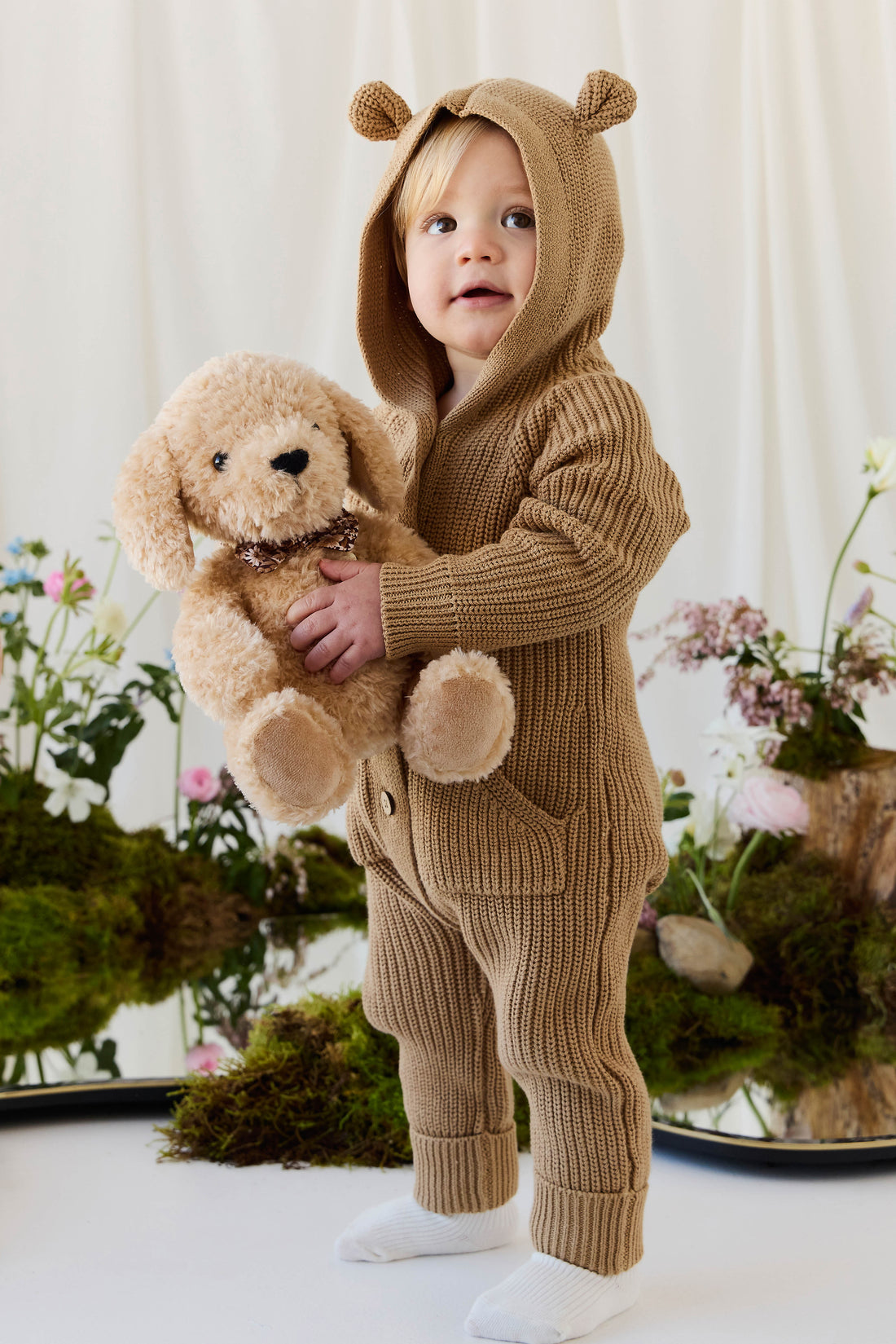 An eye-level studio portrait of a toddler standing proudly indoors against a light-colored backdrop, wearing a matching textured brown knit romper with an attached hood featuring small, upright bear ears and a front kangaroo pocket. The toddler is holding a plush honey-toned puppy toy with floppy ears and a checkered ribbon collar against their chest with both hands. The soft, natural lighting illuminates the scene, which is styled with delicate floral arrangements and moss at the base.
