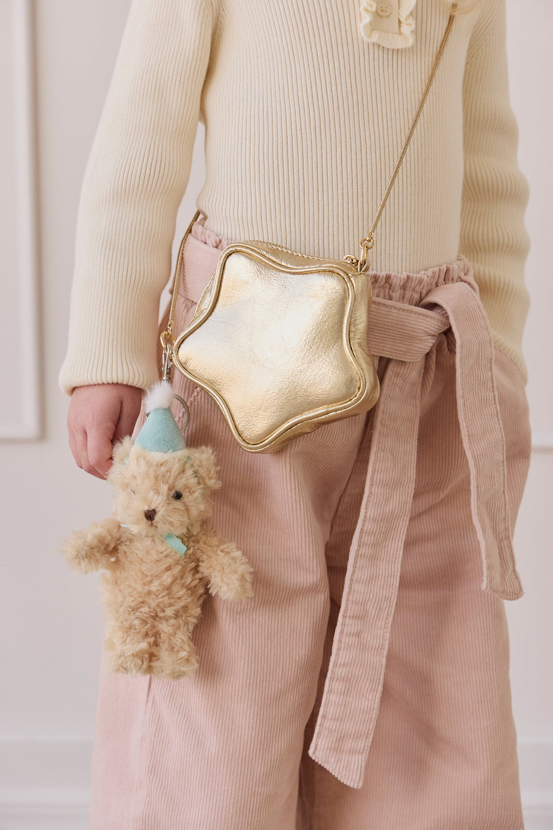 A young girl with blonde hair and a cream-colored knit top holding a gold star-shaped bag and the fluffy beige teddy bear keyring wearing a light blue party hat and matching ribbon.