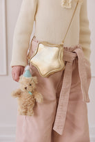 A young girl with blonde hair and a cream-colored knit top holding a gold star-shaped bag and the fluffy beige teddy bear keyring wearing a light blue party hat and matching ribbon.