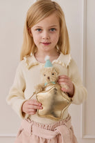 A young girl with blonde hair and a cream-colored knit top holding a gold star-shaped bag and the fluffy beige teddy bear keyring wearing a light blue party hat and matching ribbon.