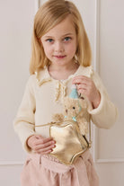 A young girl with blonde hair and a cream-colored knit top holding a gold star-shaped bag and the fluffy beige teddy bear keyring wearing a light blue party hat and matching ribbon.
