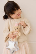 Child wearing a textured cream sweater and a pale pink tulle skirt, holding the Snuggle Bunnies Arthur Keyring by Jamie Kay attached to a metallic silver star-shaped handbag.