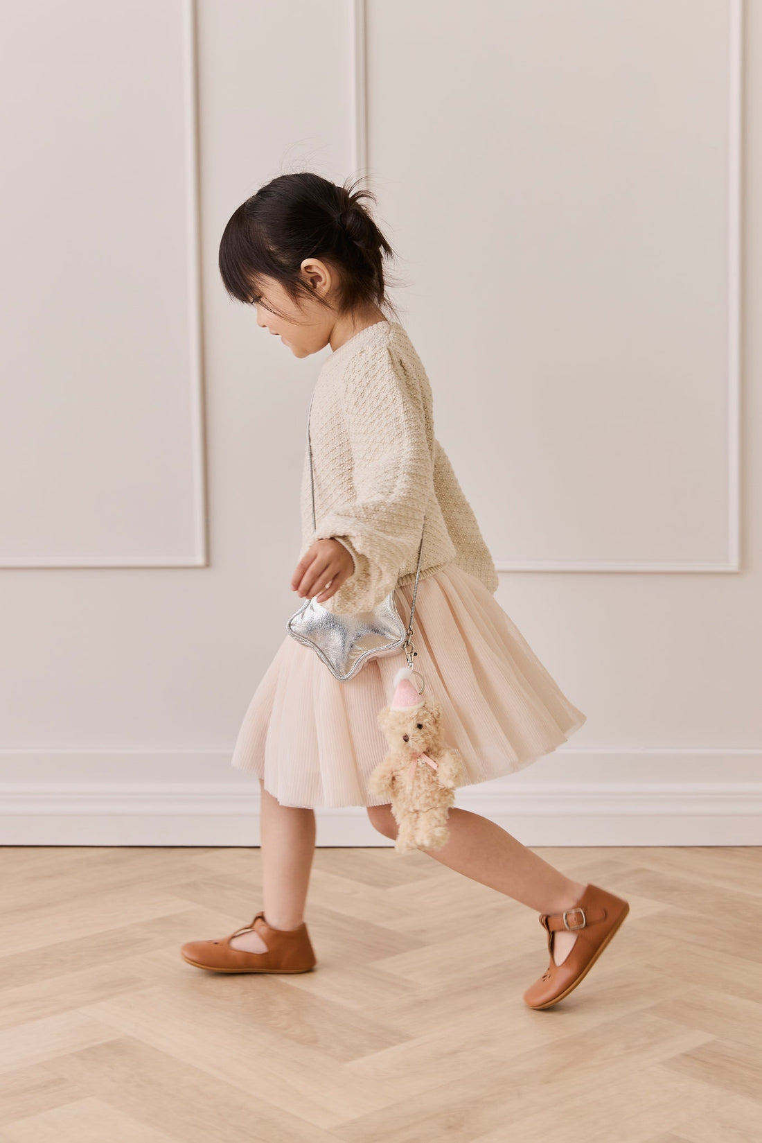 Child walking in profile view, wearing a textured cream sweater, a pale pink tulle skirt, and brown shoes, carrying a metallic silver star-shaped handbag with the Snuggle Bunnies Arthur Keyring by Jamie Kay clipped to it.