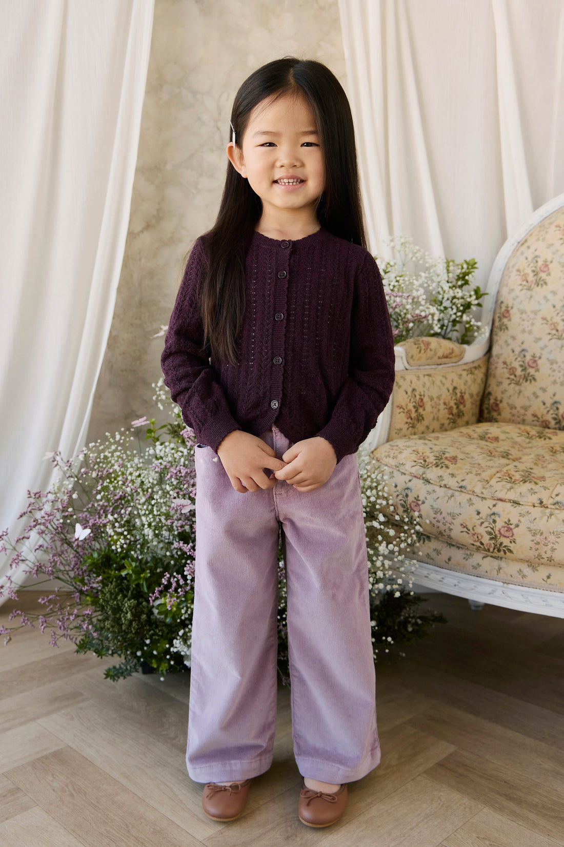 Young girl in a dark purple cardigan and light purple pants standing in a decorated room with flowers and a chair.
