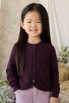 Young girl wearing a dark purple cardigan in a softly lit room with flowers and a chair.