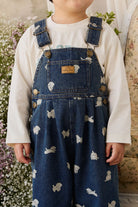 Child wearing blue denim overalls with white patterns, standing in a garden.