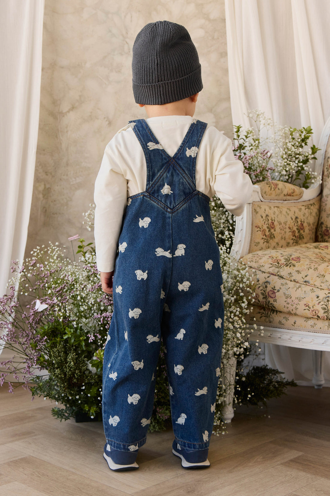 Child wearing blue overalls with white patterns, standing in a room with floral decorations and a sofa.