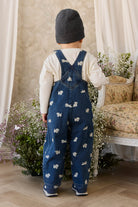 Child wearing blue overalls with white patterns, standing in a room with floral decorations and a sofa.