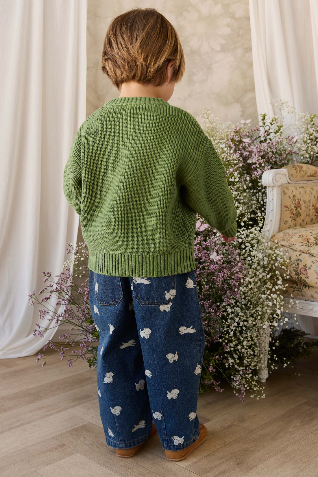 Child wearing a green sweater and blue patterned pants standing in a room with flowers and a chair.
