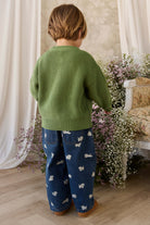 Child wearing a green sweater and blue patterned pants standing in a room with flowers and a chair.
