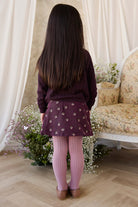 Person wearing a purple sweater and skirt standing in a room with floral decor and flowers.