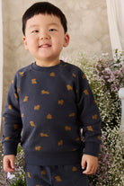 A young boy wearing a navy blue sweatshirt and matching pants set, both decorated with small mustard-gold dog silhouettes, standing in front of a soft-focus floral background.