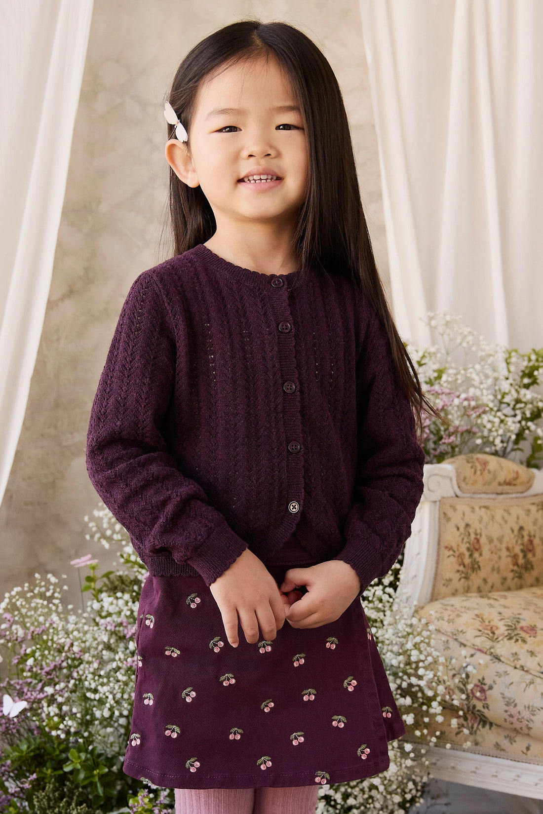 A young girl with long dark hair wearing a dark purple cable-knit cardigan and a matching purple skirt with small floral prints, standing in a decorated indoor setting with flowers and a vintage-style chair.