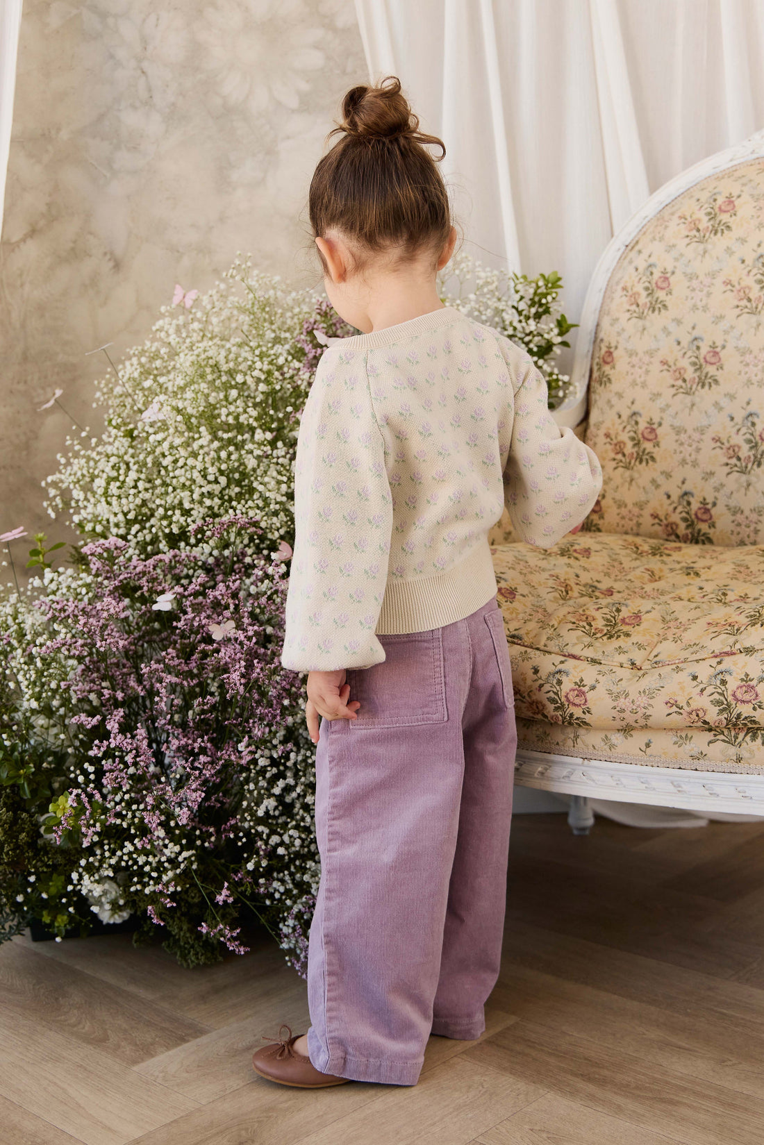 Child in a white sweater and purple pants standing near flowers and a floral-patterned couch.