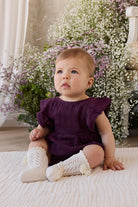 Baby in a purple outfit with white booties sitting on a blanket with flowers in the background