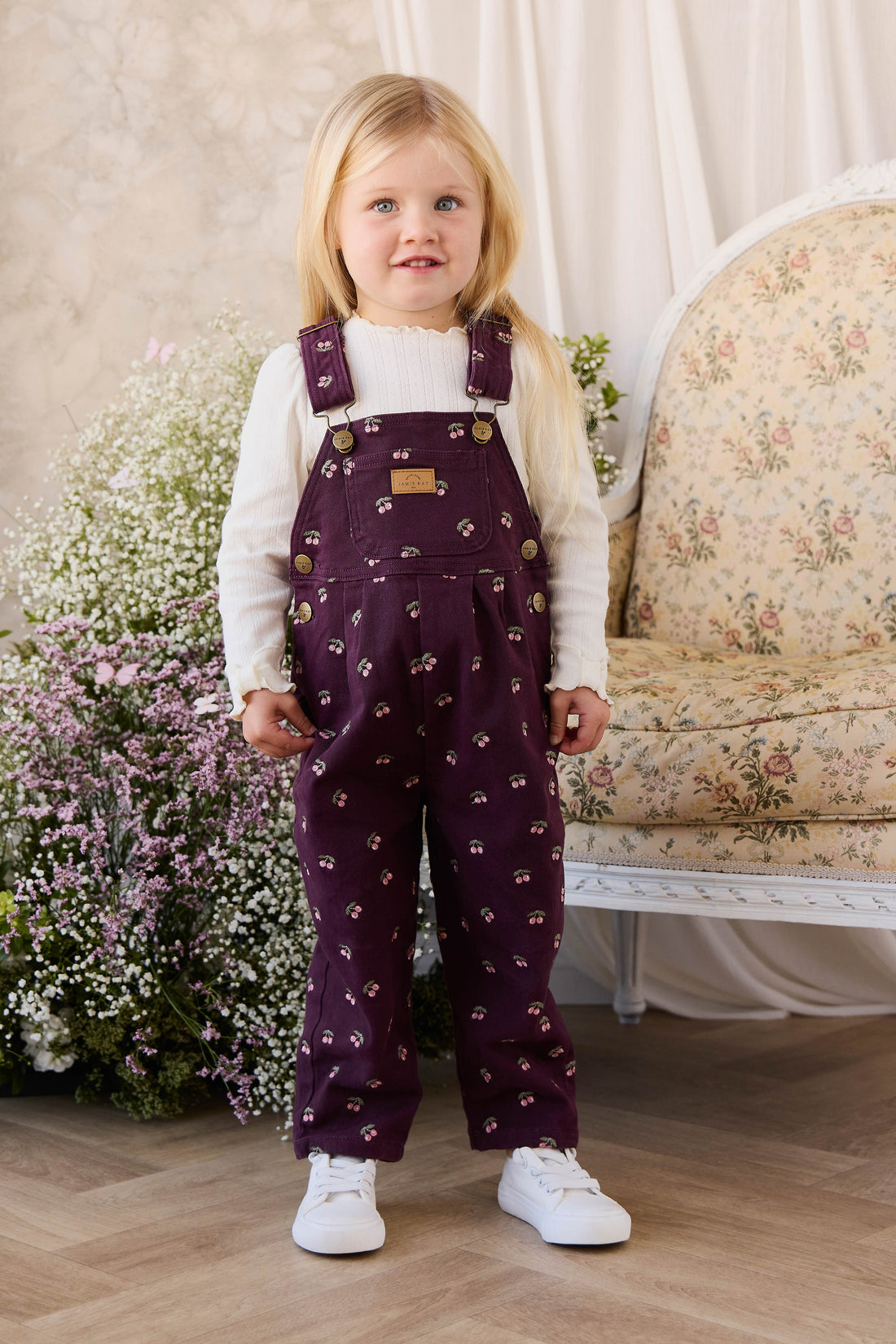 Young girl wearing a white shirt and purple floral overalls standing in a room with flowers and a floral-patterned couch.