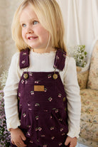 Young girl wearing a purple floral dress over a white shirt, standing in a softly lit room.