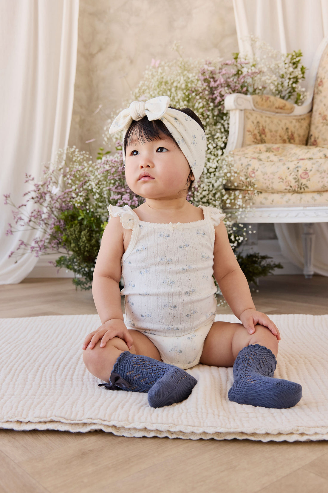 Baby sitting on a blanket wearing a white outfit, blue socks, and a headband with flowers.